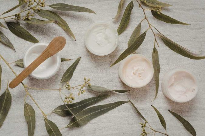 Routine bien-être avec pots de crèmes naturelles blanches et roses, spatule en bois et branches d'eucalyptus sur fond de lin.