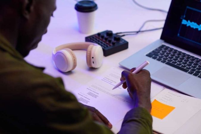 Un créateur de contenu prépare le script de son émission à son bureau avec un ordinateur affichant un logiciel audio.