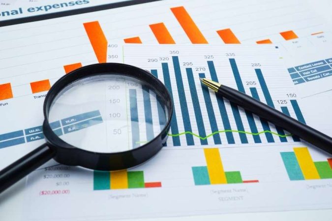 Une loupe et un stylo noir posés sur des documents comptables comportant plusieurs graphiques à barres colorés.