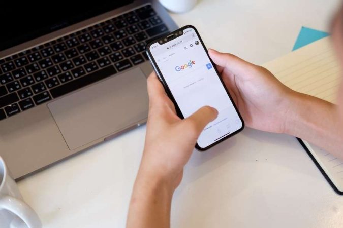 Une personne utilise son smartphone pour afficher la page d'accueil de recherche Google à côté d'un ordinateur portable.