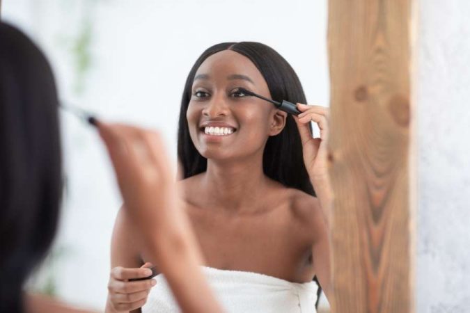 Une femme noire se maquille les yeux avec du mascara devant un miroir.