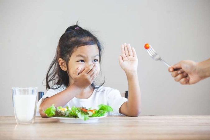 Une petite fille refuse de manger des légumes en faisant un geste de la main.