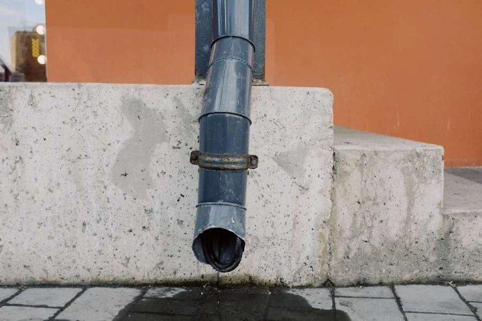 Un tuyau de descente d'eau grise extérieur en métal gris face à un mur orange, souvent lié aux problèmes de mauvaises odeurs de canalisation.